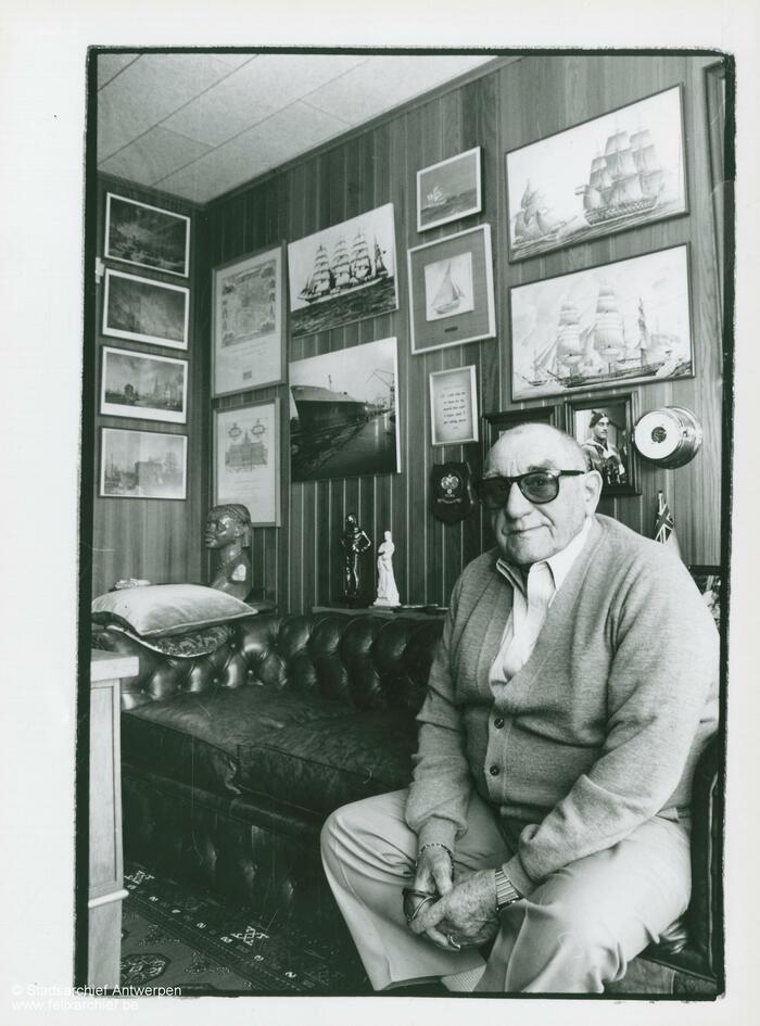 Eugène Colson on a sofa, with a wall with frames behind him
