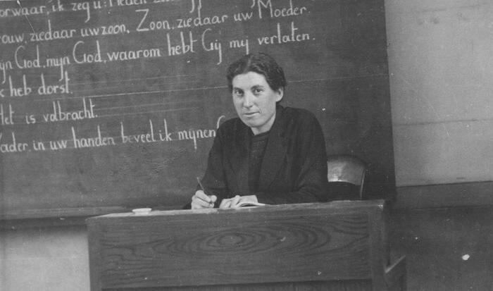 A woman sits behind a desk in front of a blackboard